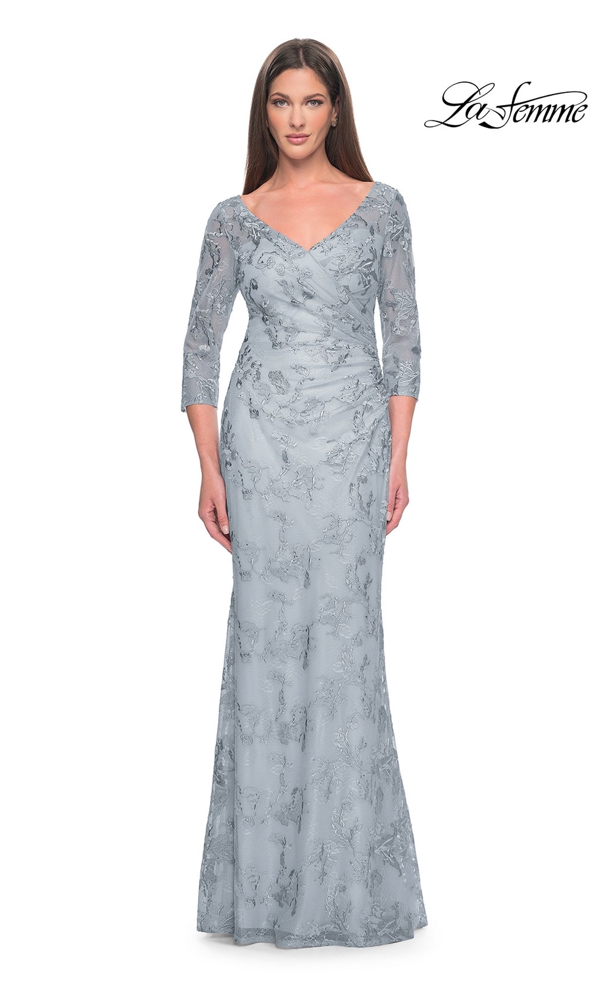 A woman in the La Femme Embroidered Lace Pastel Formal Dress 31684 in dusty blue, featuring sheer three-quarter sleeves, a V-neckline, floor-length skirt, and delicate floral embroidered lace, stands against a white background.